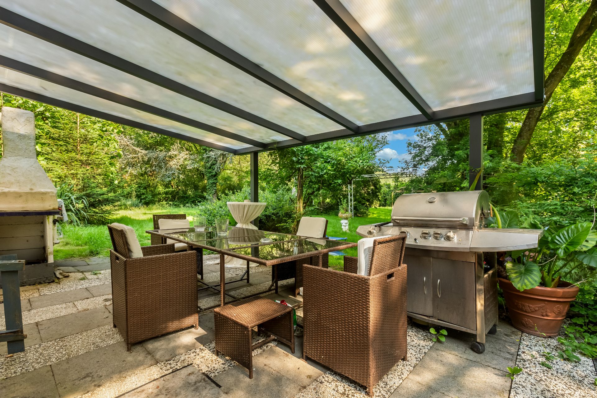 Überdachte Terrasse mit Glas-Esstisch, Stühlen, Grill und Blick auf einen grünen Garten.