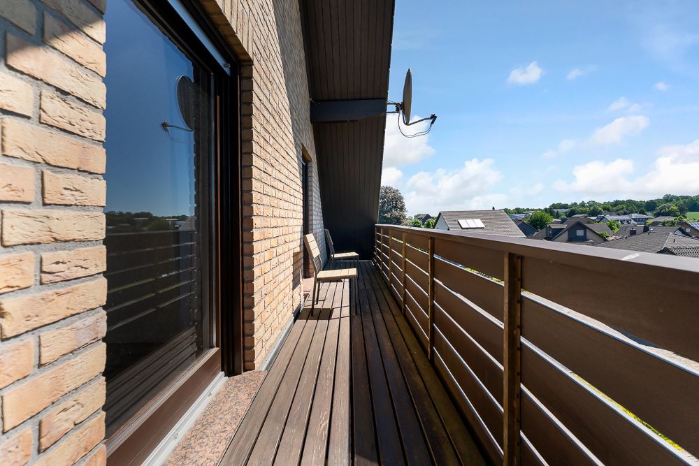 Langer Balkon mit Holzboden, Stühlen und Blick auf Dächer und grüne Landschaft.