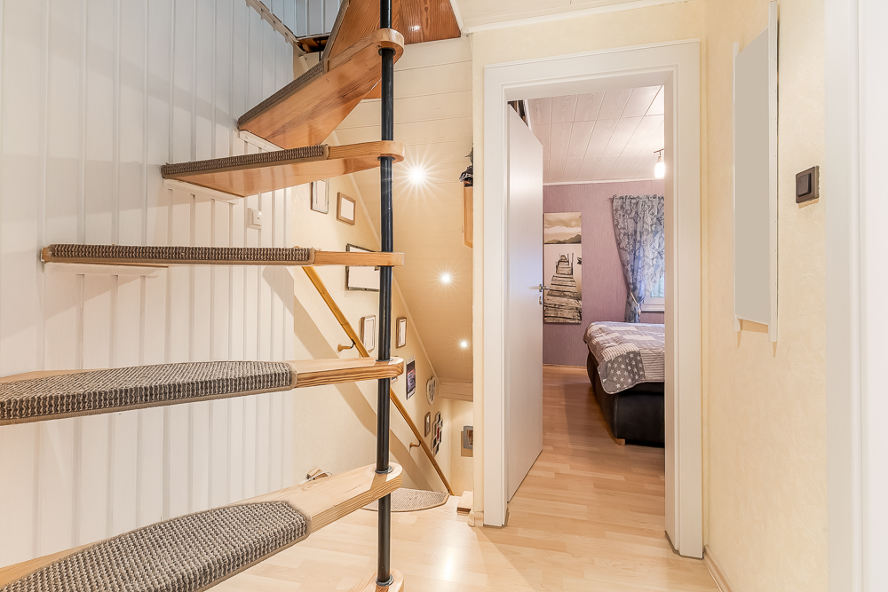 Holztreppe mit Teppichstufen, Flur und Blick in ein Schlafzimmer mit Bett und Fenster.