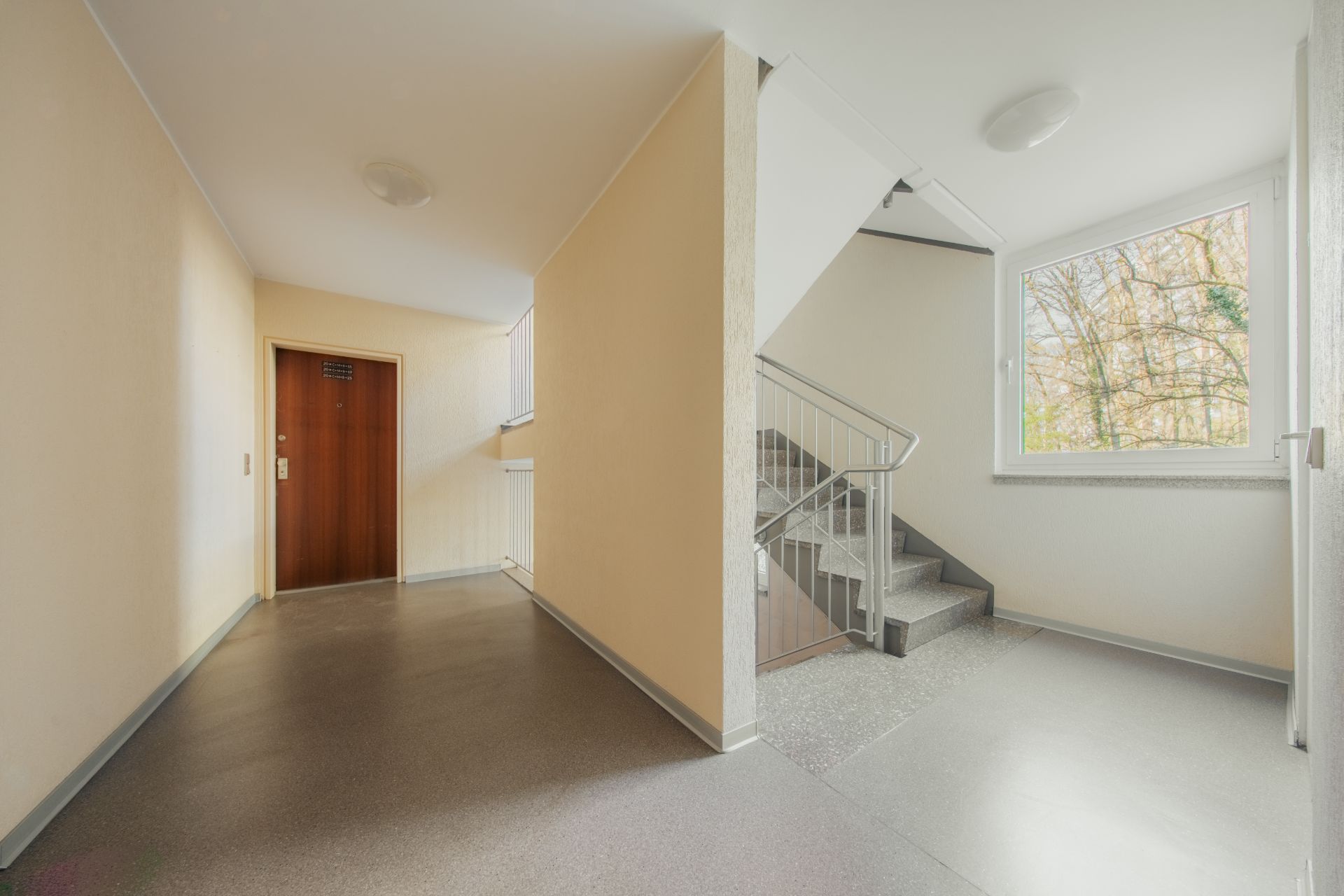 Heller Hausflur mit Treppe, großem Fenster und brauner Wohnungstür.