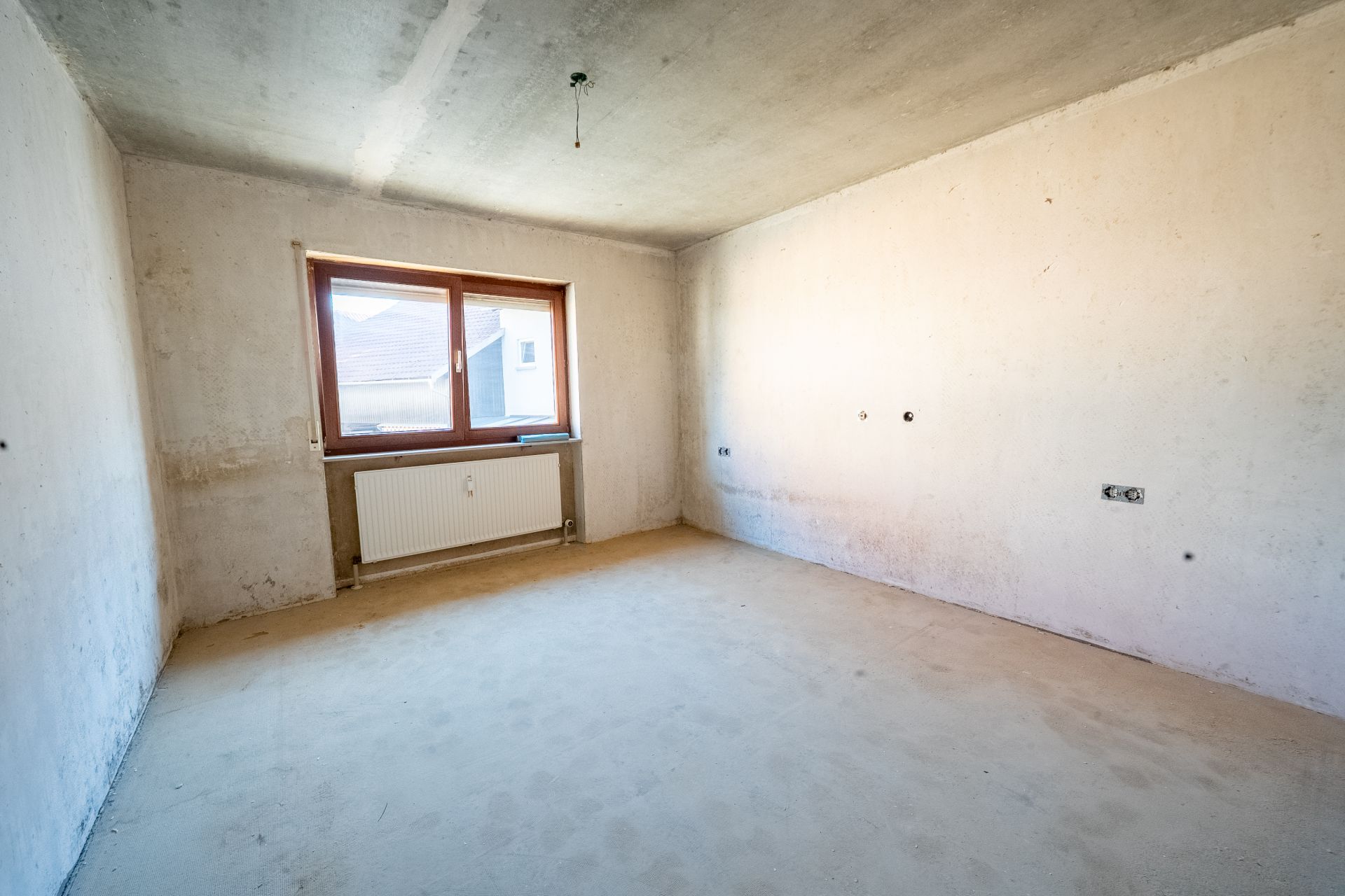 Leerer Raum mit unverputzten Wänden, einem Fenster und einem Heizkörper unter dem Fenster.
