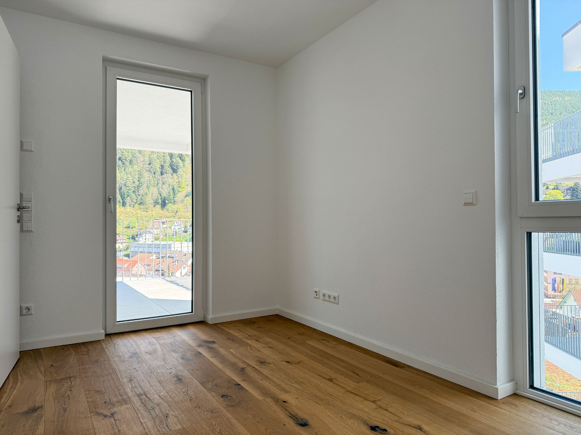 Leerer, heller Raum mit Holzboden und bodentiefen Fenstern, Blick auf Balkon und grüne Landschaft.