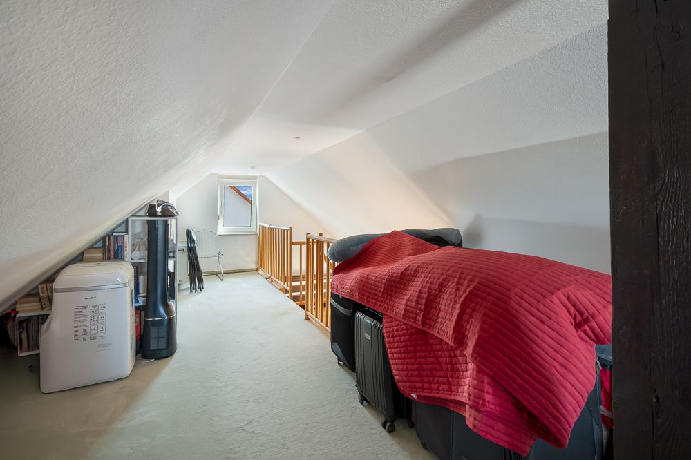 Dachzimmer mit Bett, roter Decke, Holzgeländer und kleinem Fenster.