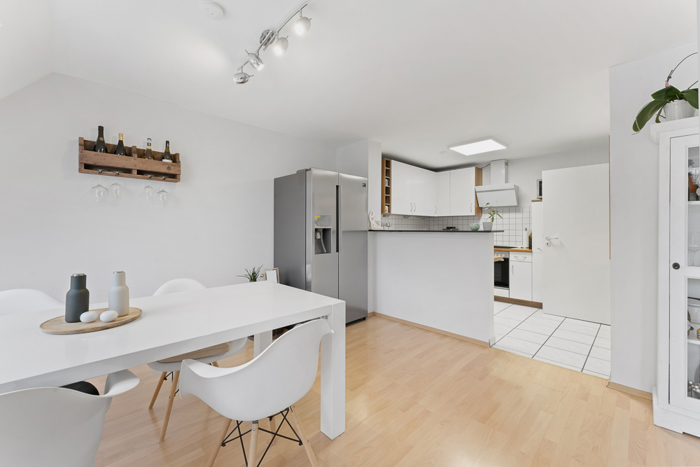 Helle Küche mit Esstisch, weißen Stühlen, großem Kühlschrank und Wandregal mit Flaschen.