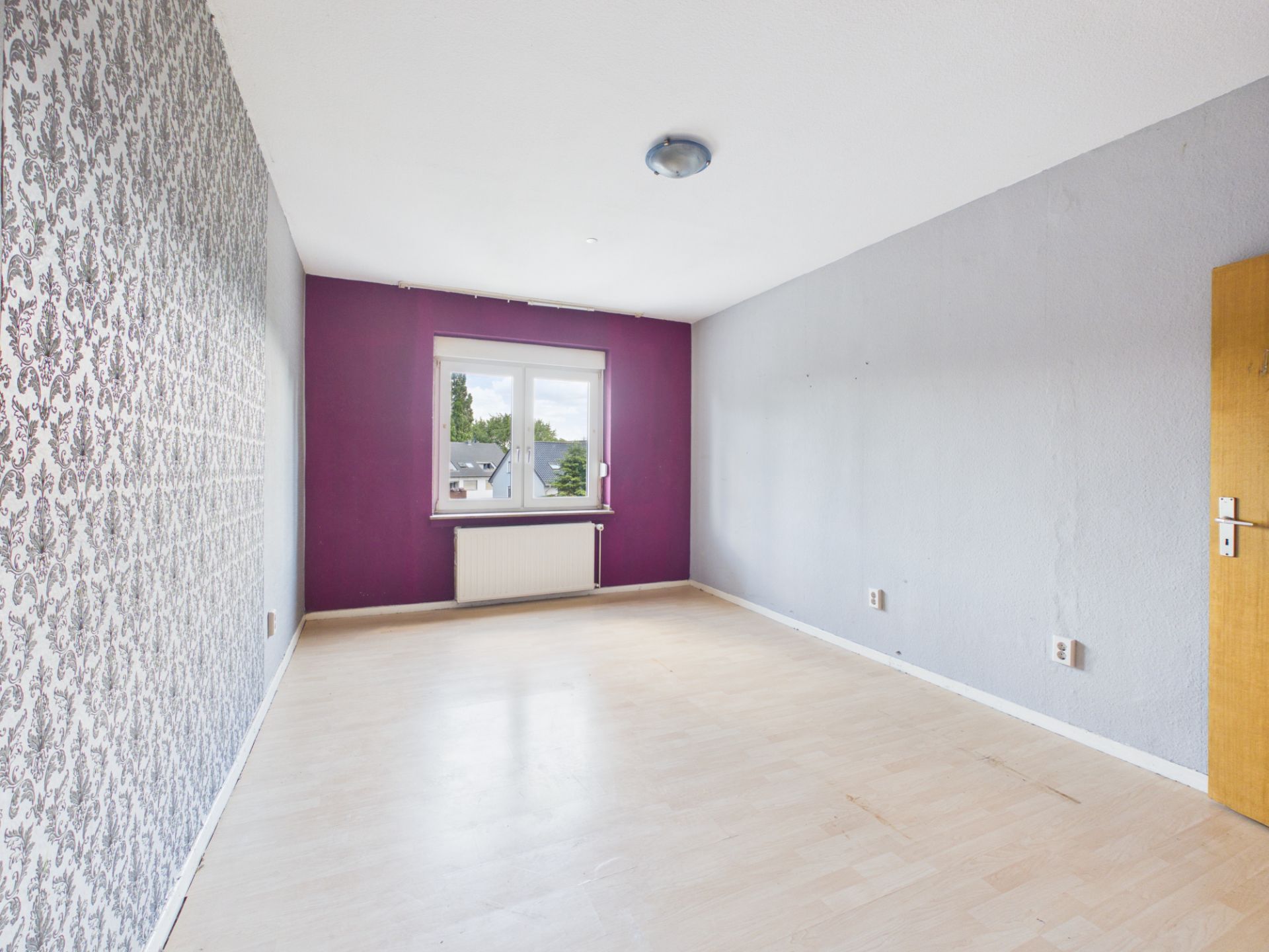 Helles, leeres Zimmer mit gemusterten und lila Wänden, Holzboden und Fenster.