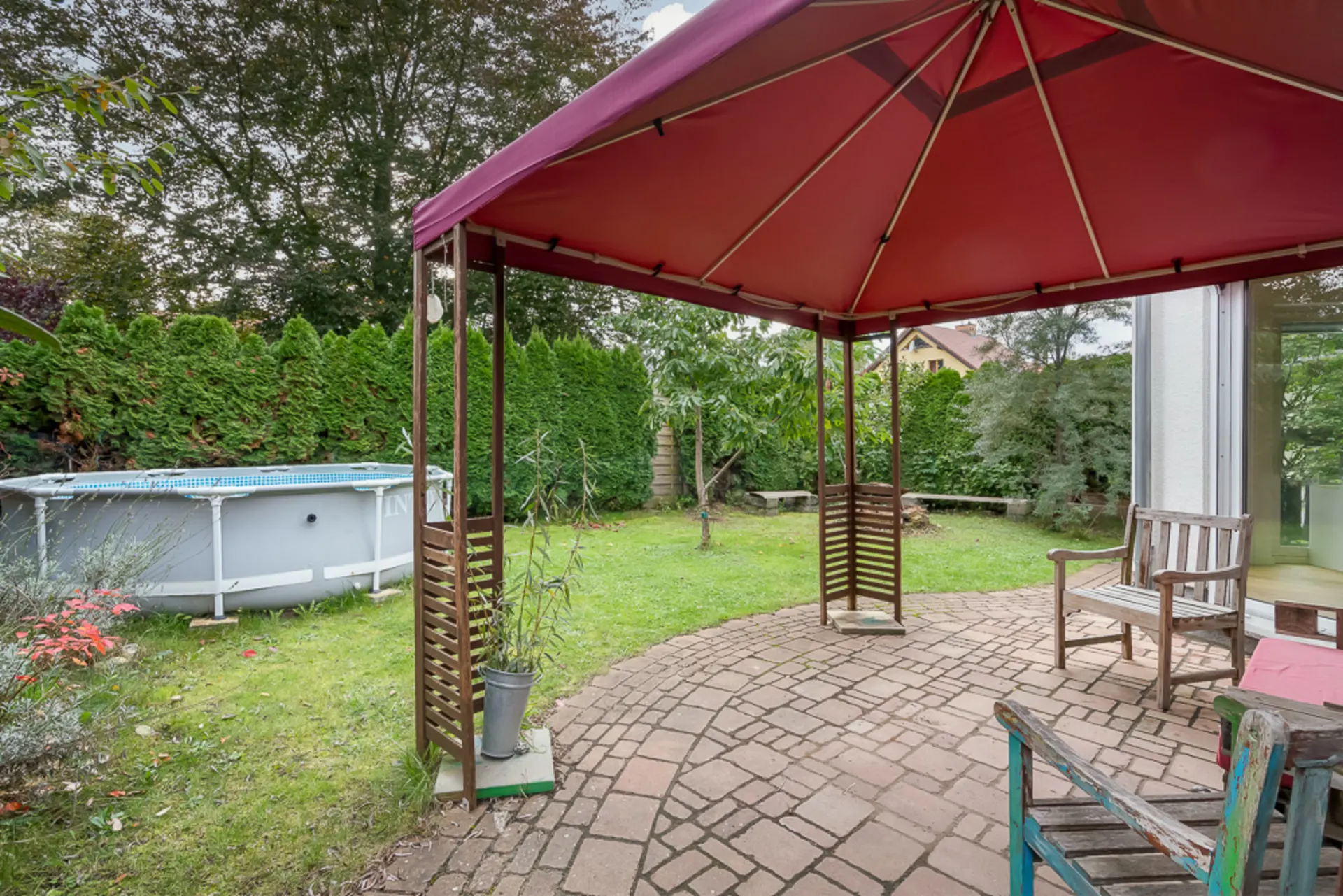 Überdachte Terrasse mit Holzmöbeln, Blick auf Garten und Aufstellpool, umgeben von Bäumen und Hecken.