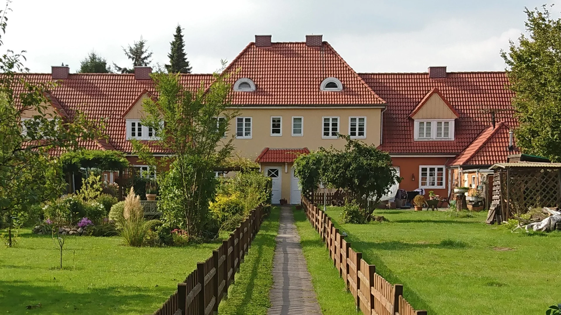 Mehrfamilienhaus mit rotem Dach, Garten und Holzzaun im Vordergrund.