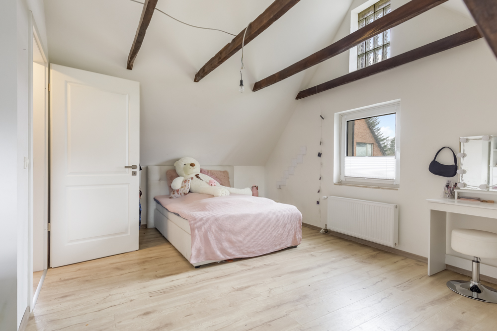 Helles Schlafzimmer mit Holzbalken, Bett, großem Teddybär, Fenster und Schminktisch.