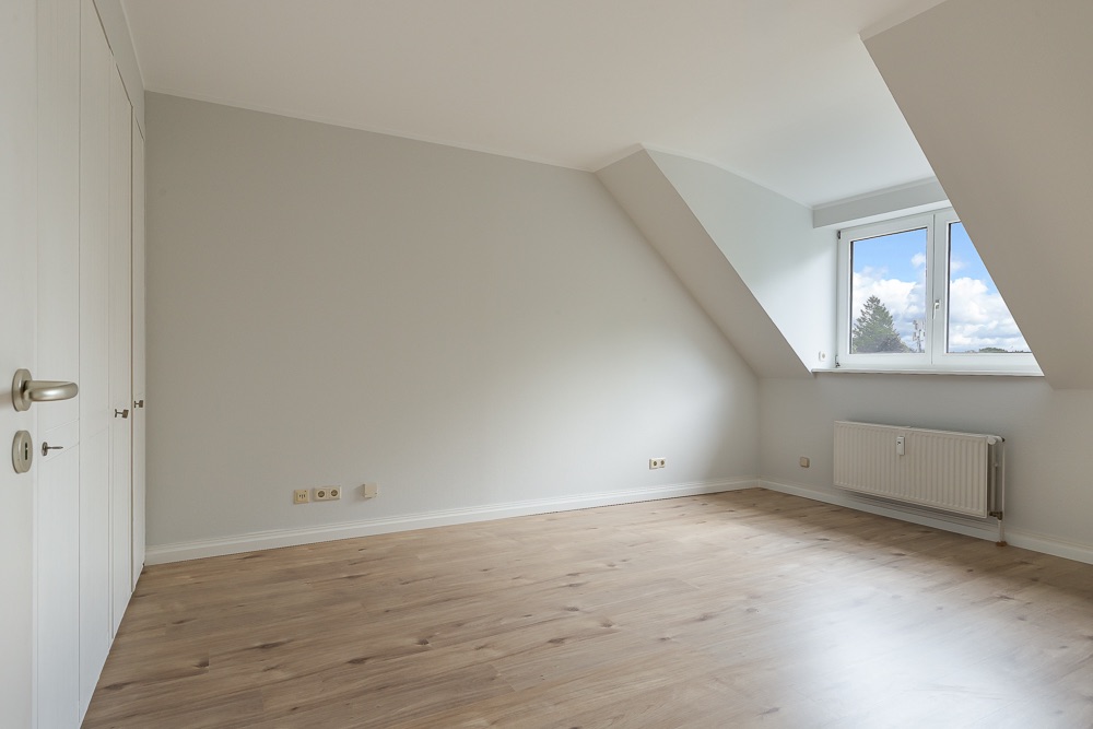 Leeres Zimmer mit hellem Holzboden, Dachschräge und Fenster mit Blick nach draußen.