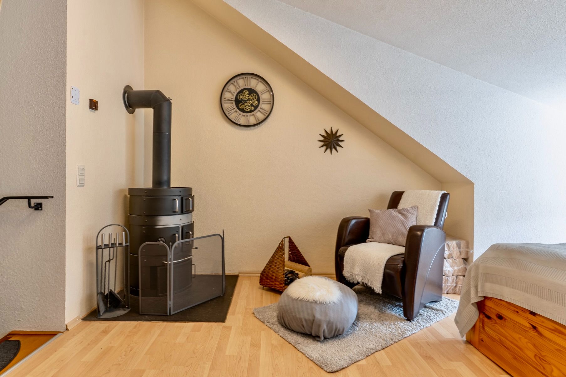 Gemütliche Ecke mit Sessel, Holzboden, Kaminofen und dekorativer Wanduhr unter Dachschräge.