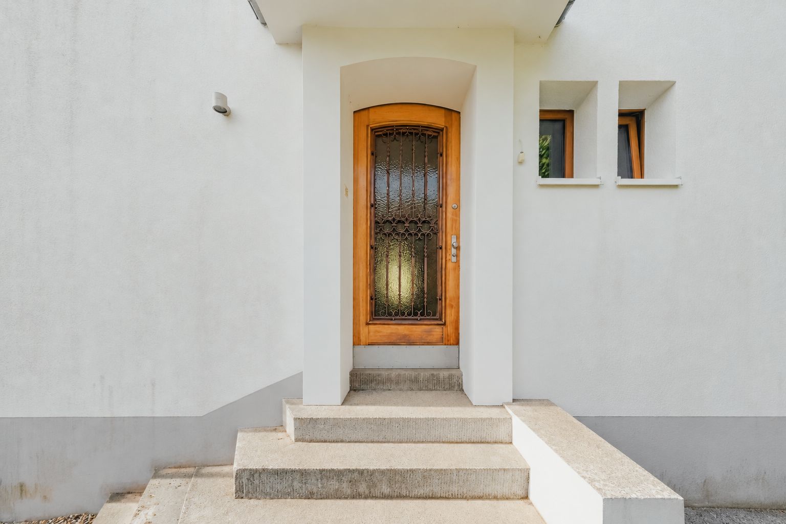Eingangstür aus Holz mit Glaseinsatz und Stufen, daneben zwei kleine Fenster.