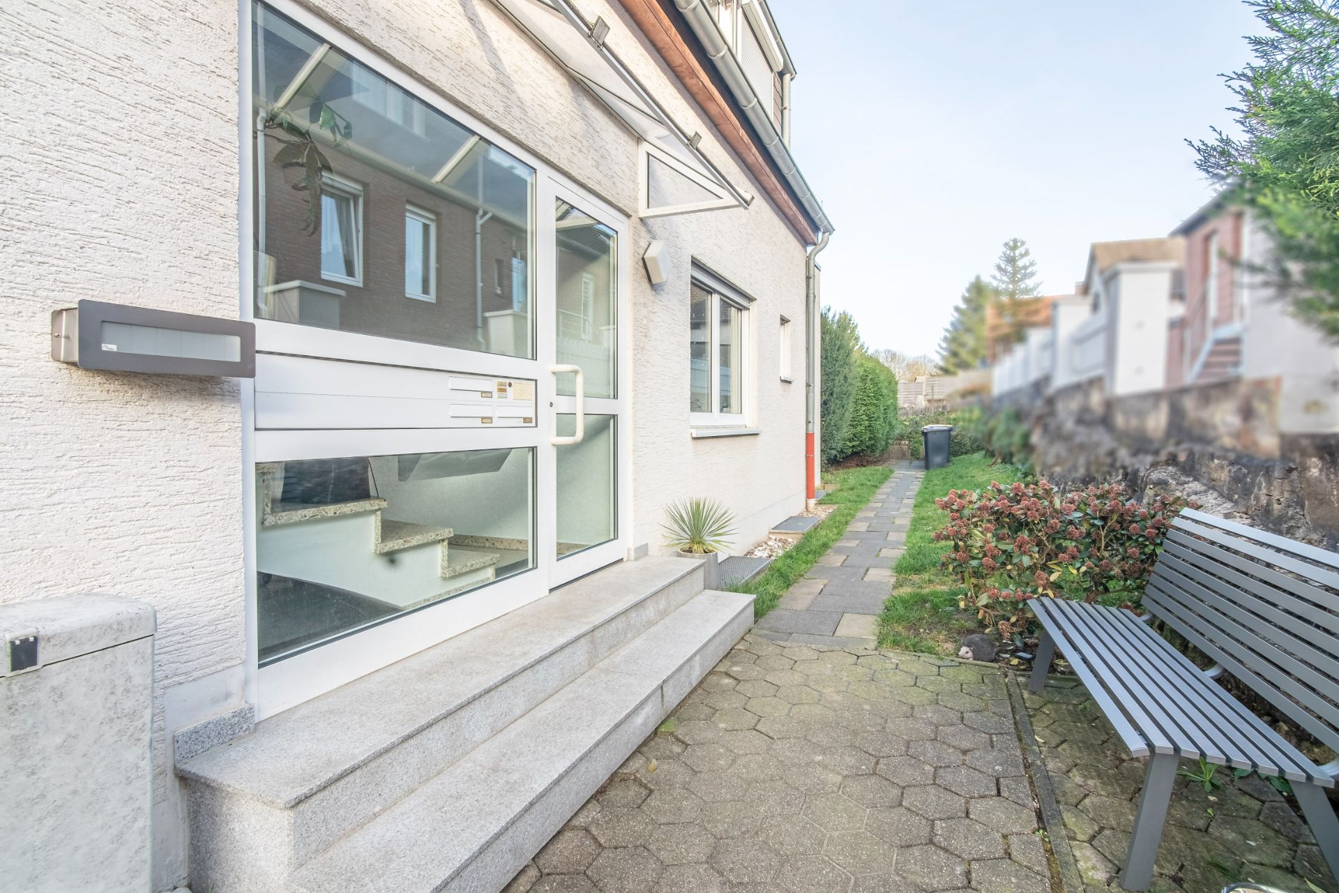 Eingang eines Wohnhauses mit Glastür, Treppen, Bank und Gehweg im Vorgarten.