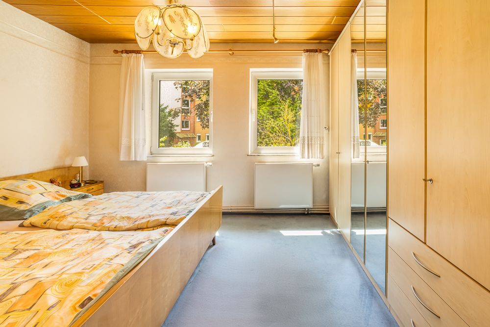 Helles Schlafzimmer mit Doppelbett, Kleiderschrank und zwei Fenstern.