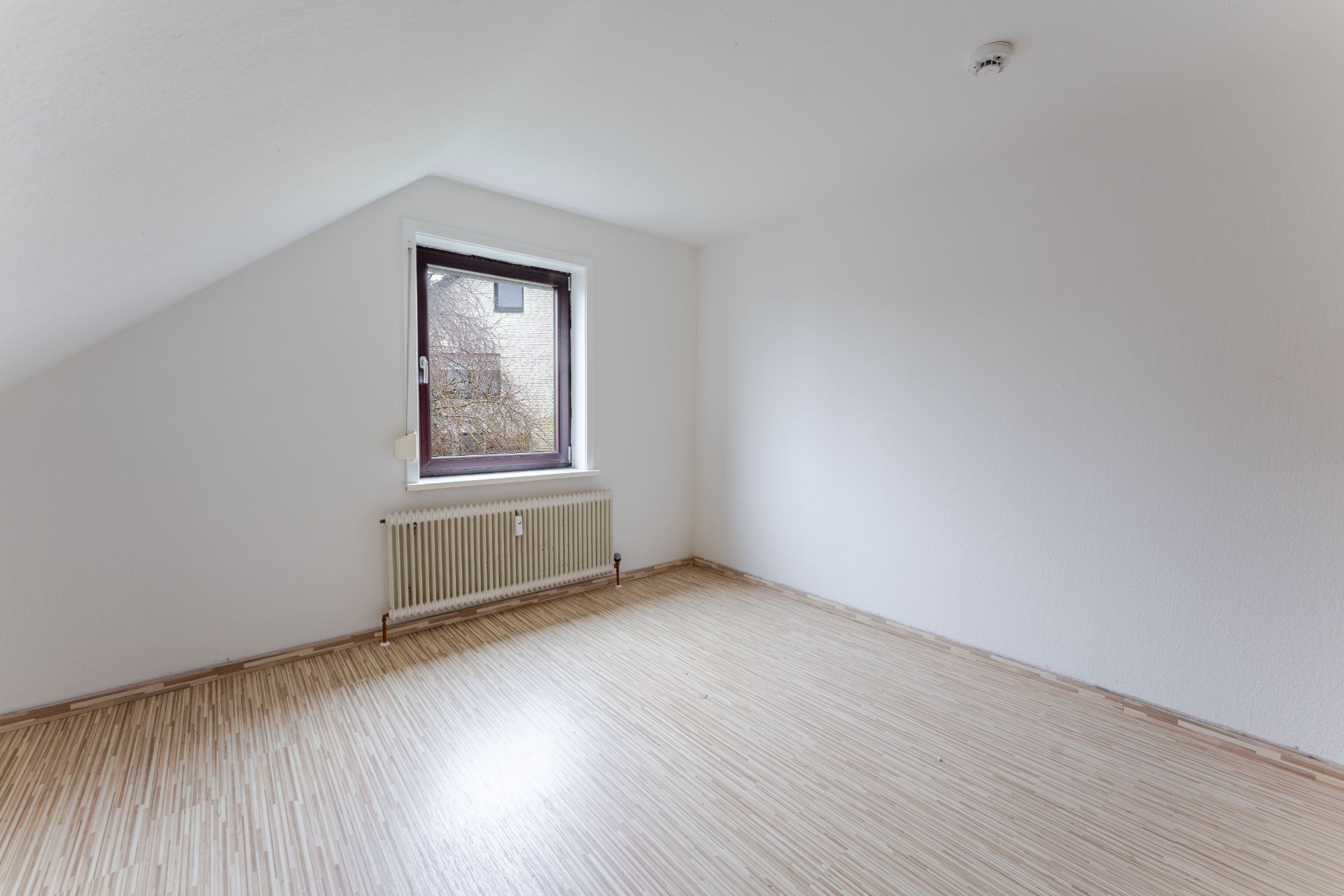 Leeres Zimmer mit Holzfußboden, weißer Wand, Fenster und Heizkörper unter dem Fenster.
