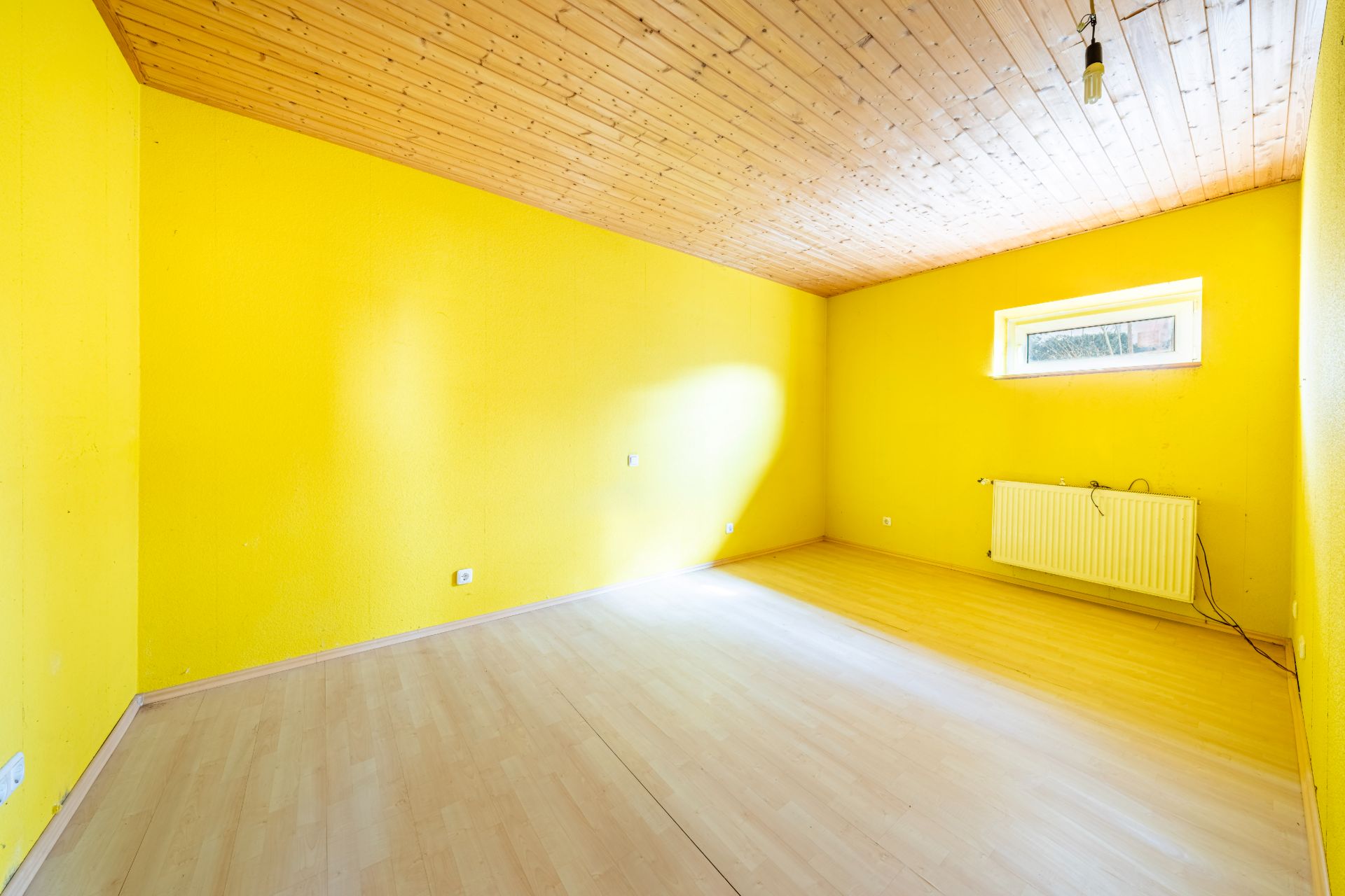 Leerer Raum mit gelben Wänden, Holzdecke, hellem Boden und kleinem Fenster.