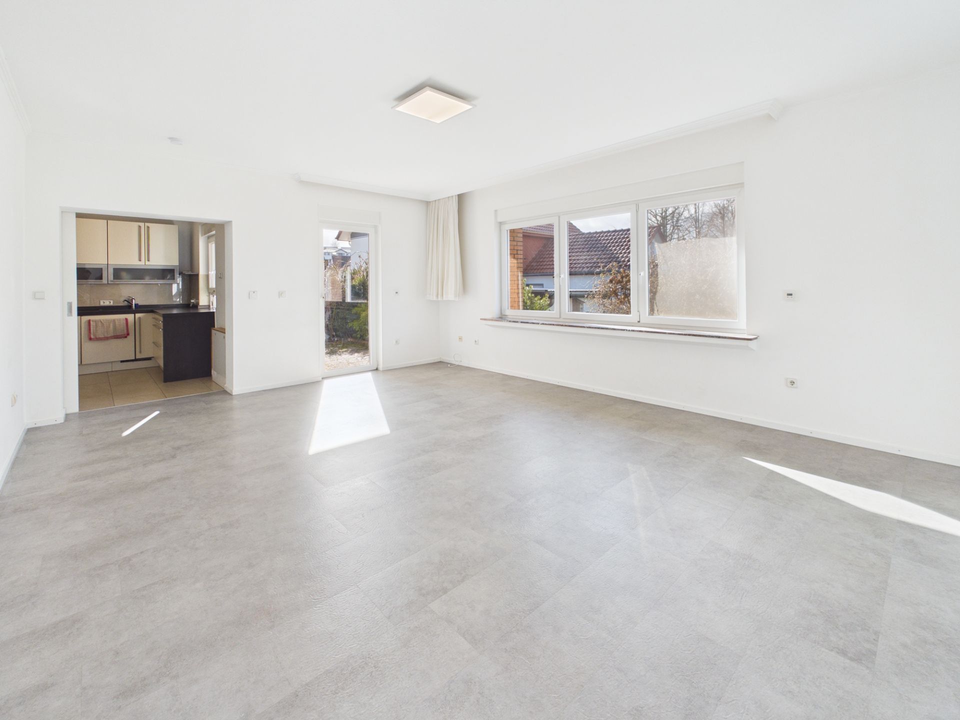 Helles, leeres Wohnzimmer mit großem Fenster, Tür zum Garten und Blick in eine Küche.