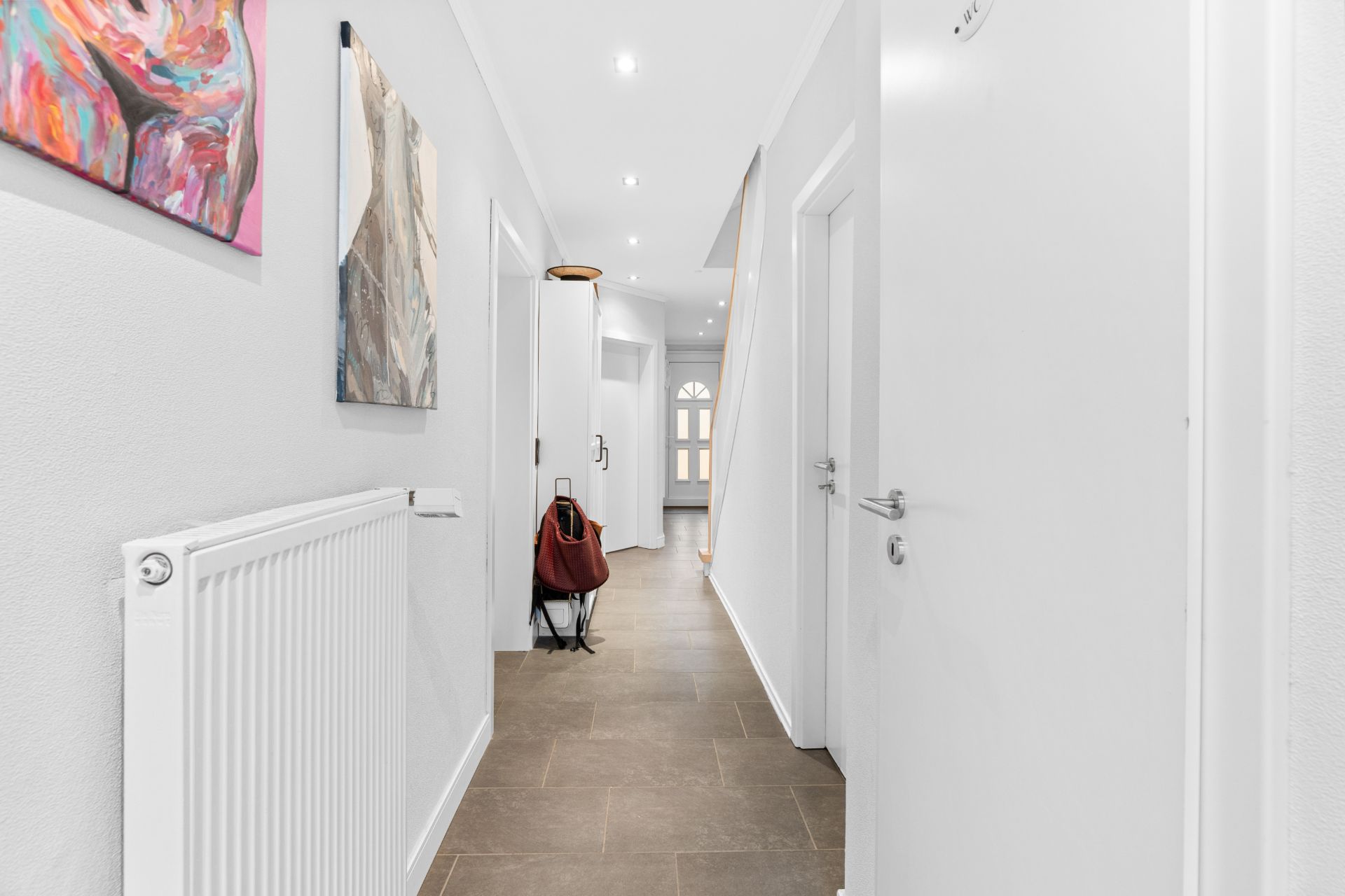 Heller Flur mit weißen Wänden, braunem Boden, Heizkörper und bunter Kunst an der Wand.