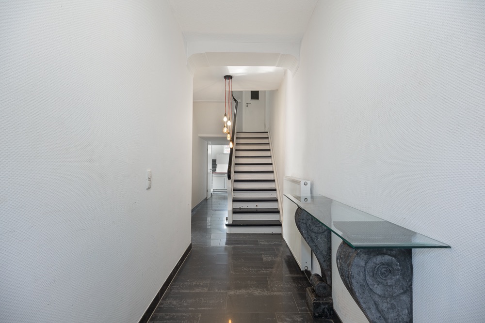Heller Flur mit dunklem Boden, Treppe am Ende und Glasablage an der rechten Wand.