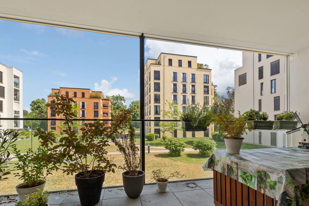 Balkon mit mehreren Blumentöpfen, Ausblick auf moderne Wohngebäude und Grünfläche.