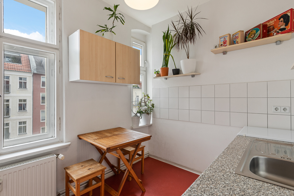 Helle Küche mit kleinem Holztisch, zwei Hockern, Pflanzen und Wandregalen vor Fenstern.