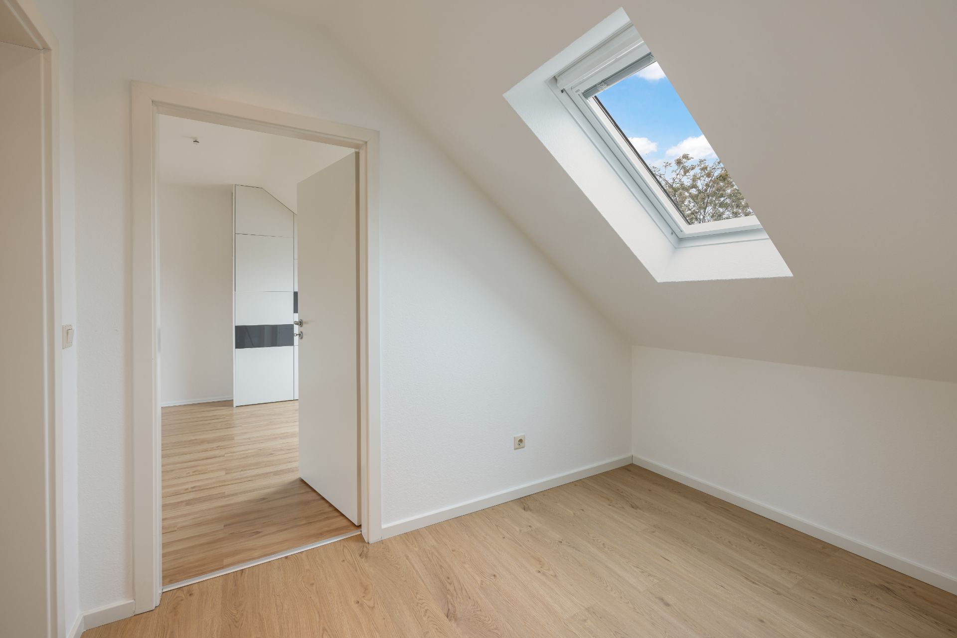 Helles Zimmer mit Dachfenster, Holzfußboden und offener Tür zu weiterem Raum.