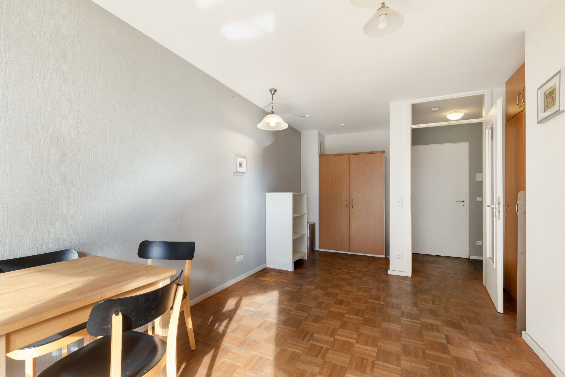 Helles Zimmer mit Holzboden, Esstisch, Stühlen, Regal und Kleiderschrank.