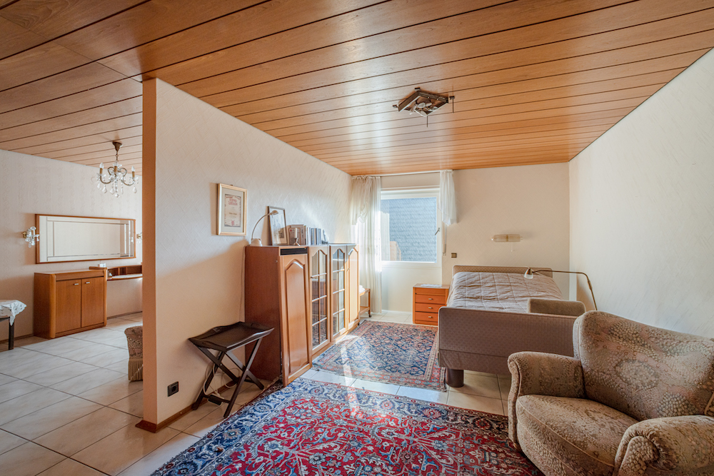 Helles Zimmer mit Einzelbett, Sessel, Teppich, Schrank und Fenster, Holzbalkendecke.