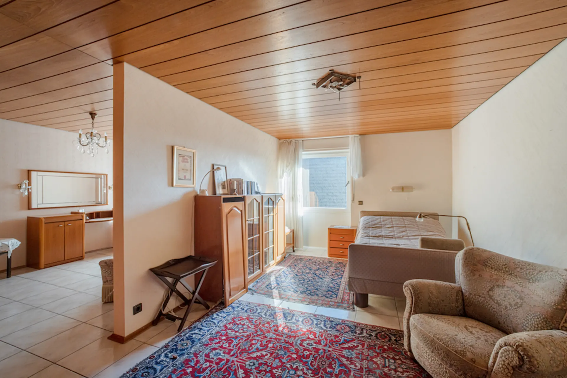 Helles Zimmer mit Einzelbett, Sessel, Teppich, Schrank und Fenster, Holzbalkendecke.