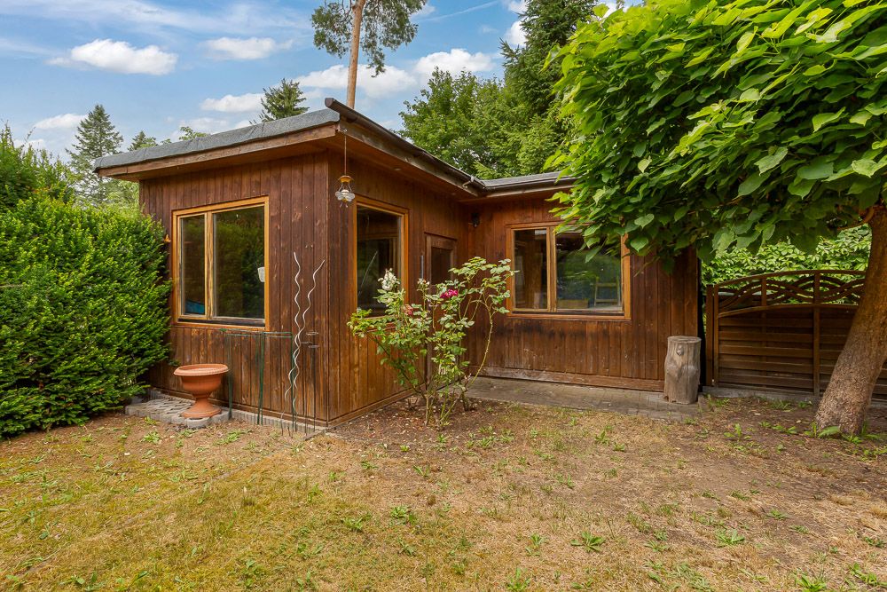 Kleines Holzhaus mit großen Fenstern im Garten, umgeben von Bäumen und Sträuchern.