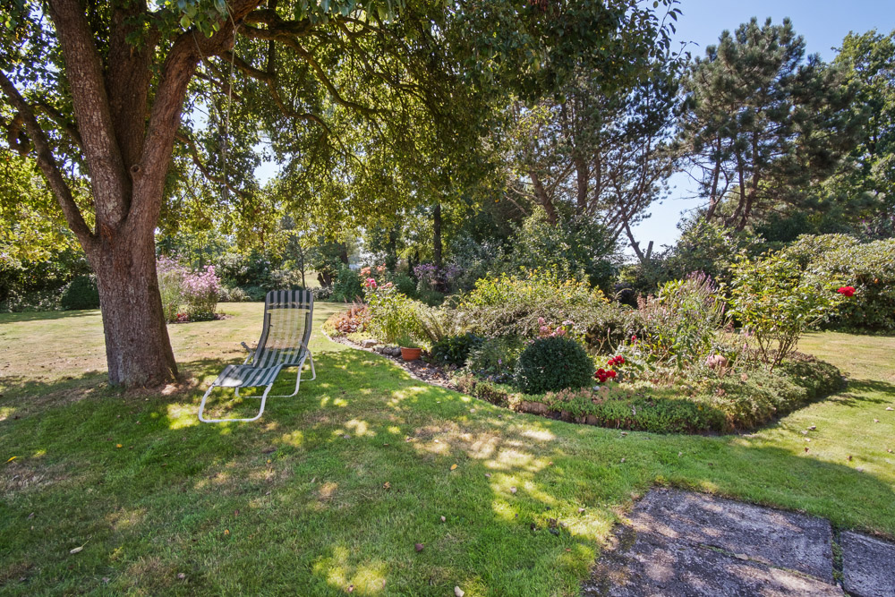 Garten mit Liegestuhl im Schatten eines Baumes und Blumenbeet im Hintergrund.