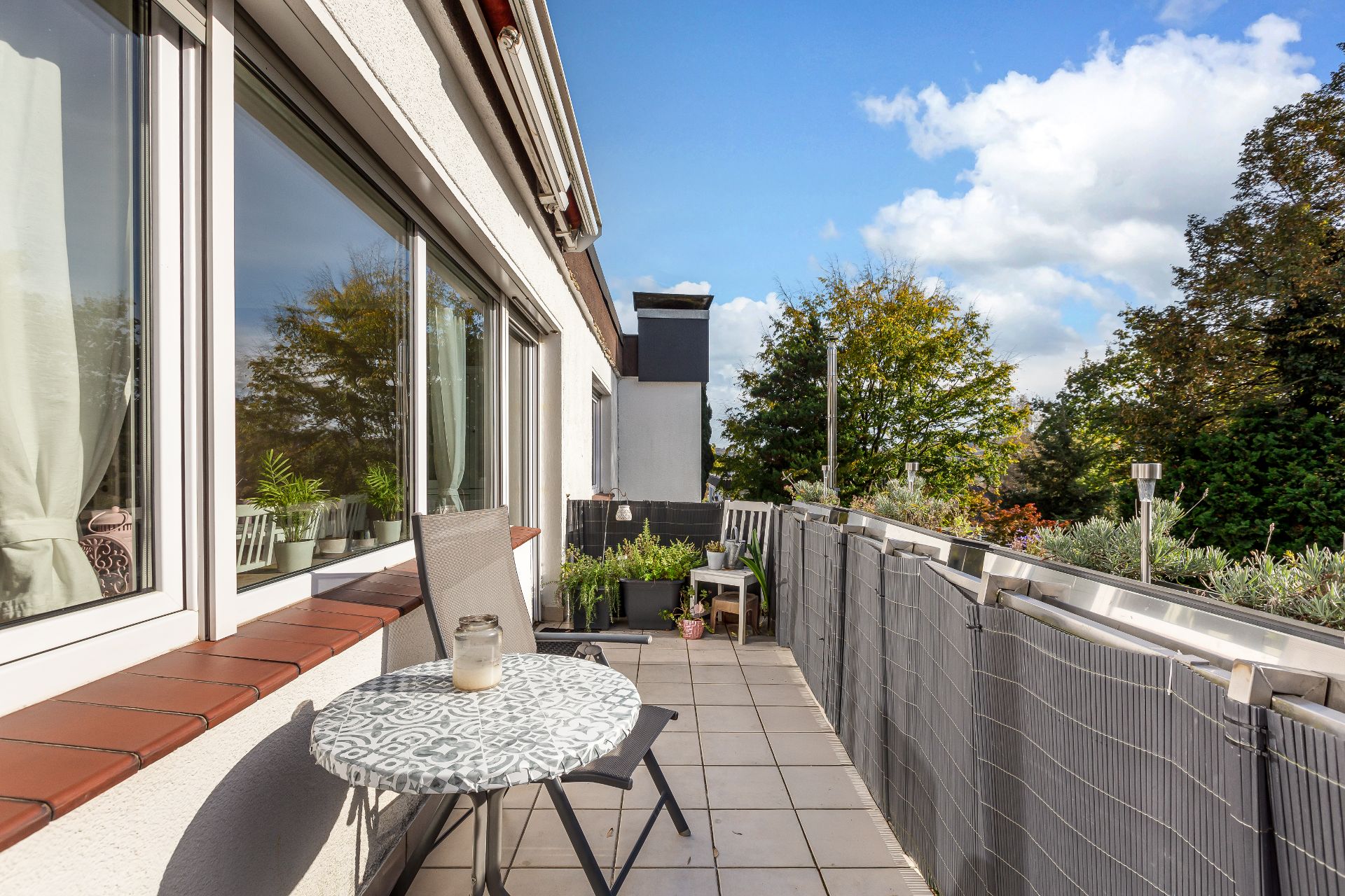 Geräumiger Balkon mit Tisch, Stuhl und Pflanzen, umgeben von Bäumen und mit Blick auf den Himmel.