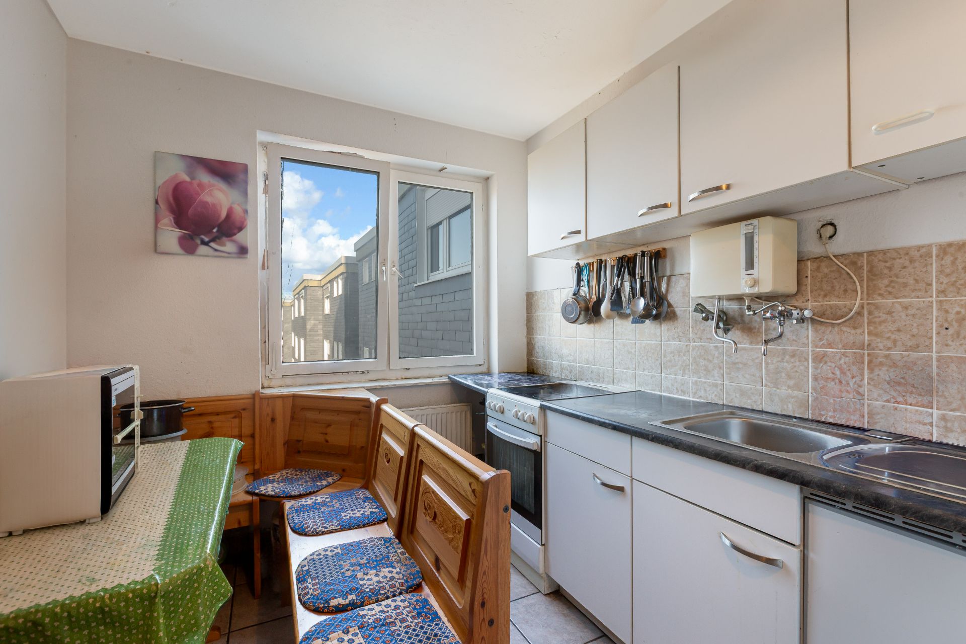 Kleine Küche mit Fenster, Holzbänken, Tisch, Herd, Spüle und Hängeschränken.