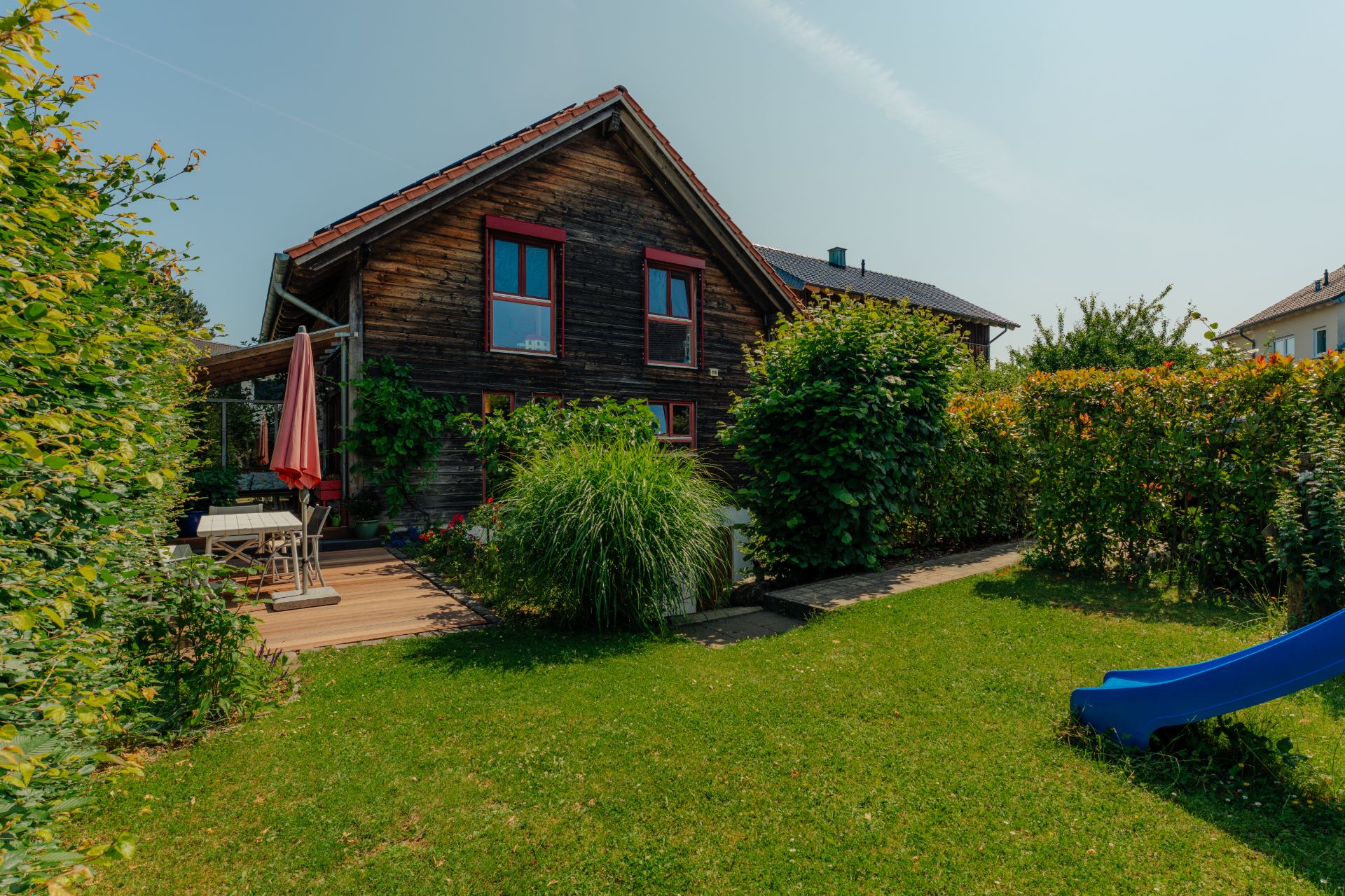 Garten mit Holzhaus, Terrasse, Tisch, Stühlen und blauer Rutsche.