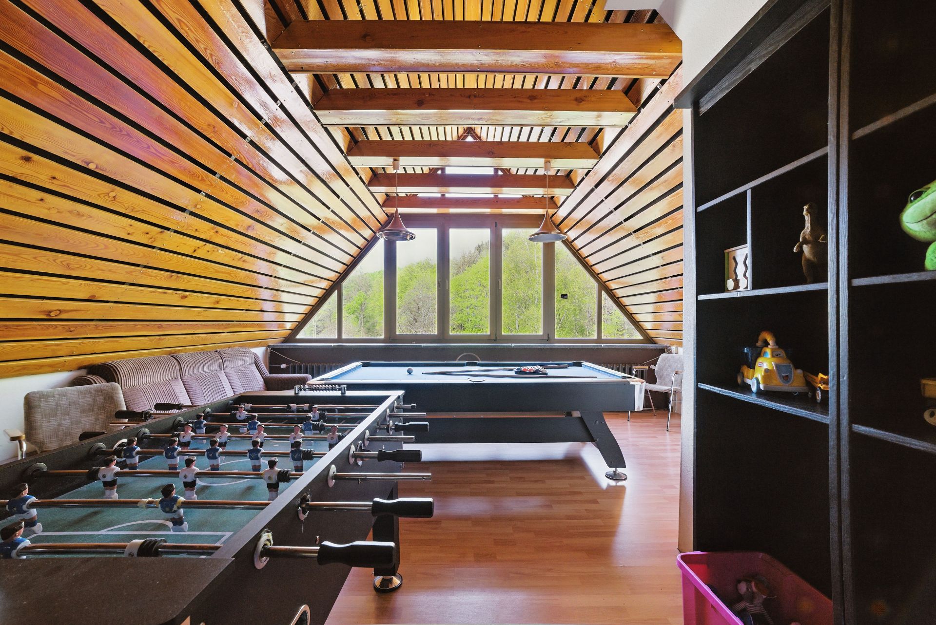 Dachzimmer mit Holzdecke, Tischkicker, Billardtisch und Regal mit Spielsachen.