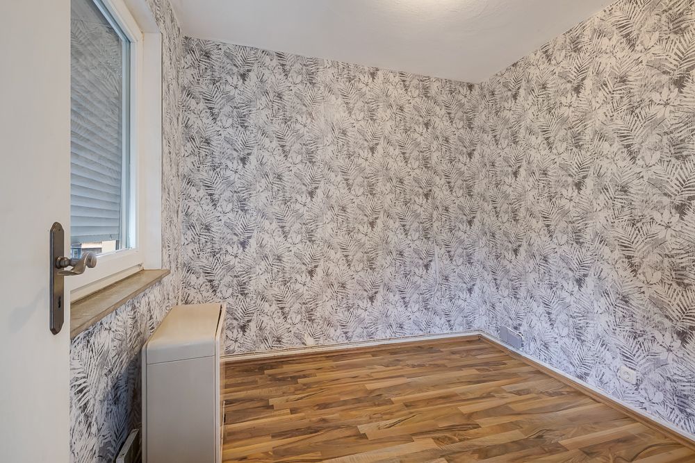 Leerer Raum mit gemusterter Tapete und Holzboden, Fenster links, Heizung unter dem Fenster.