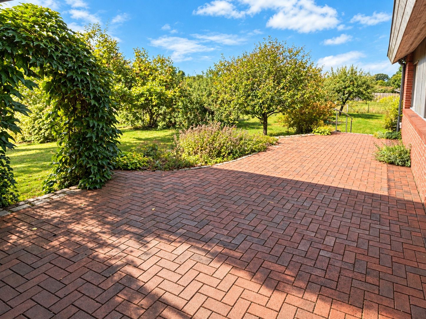 Rote gepflasterte Terrasse mit Blick auf Garten und Bäume an einem sonnigen Tag.