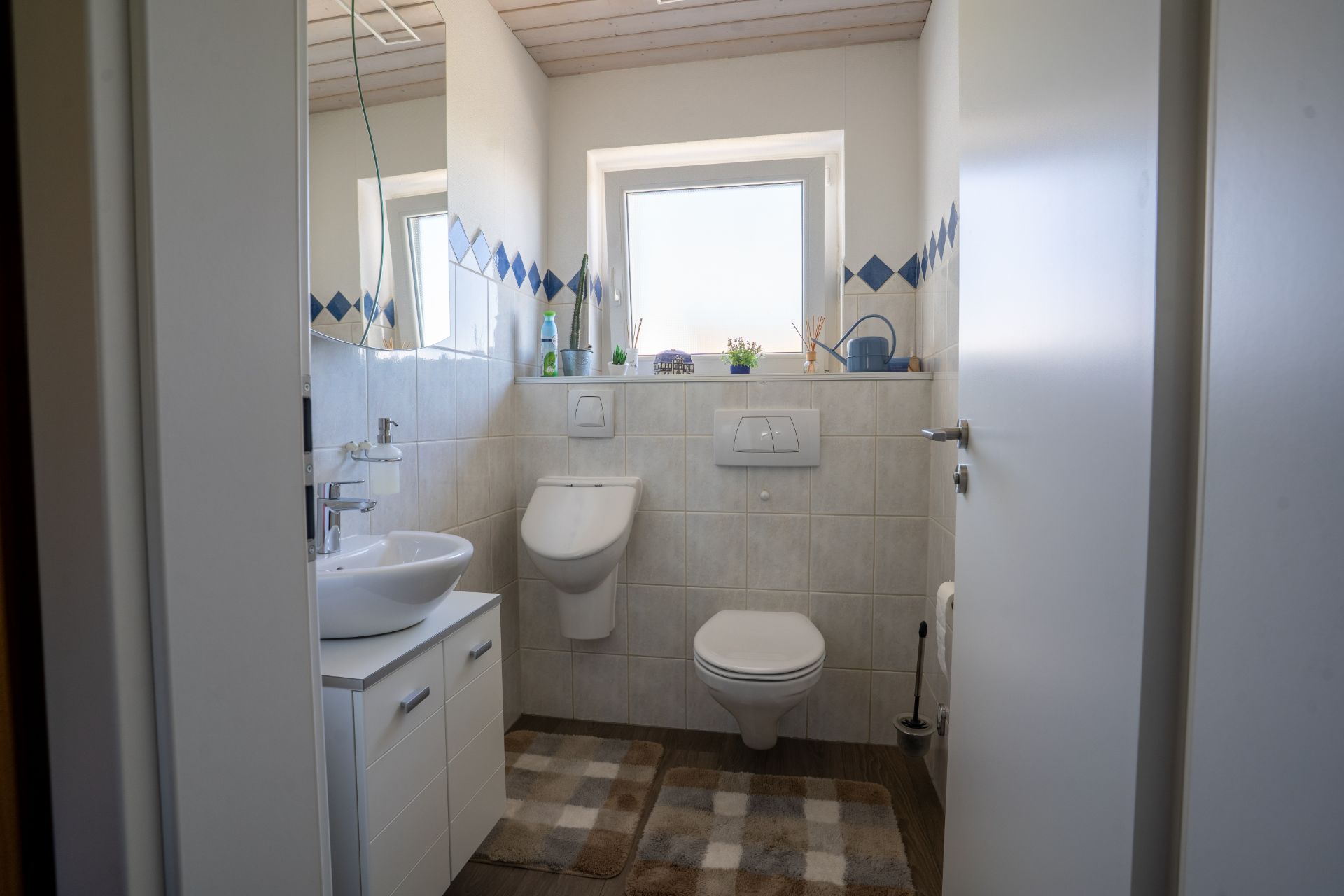 Helles Badezimmer mit Waschbecken, Urinal, Toilette, Fenster und kleinen Pflanzen auf der Fensterbank.