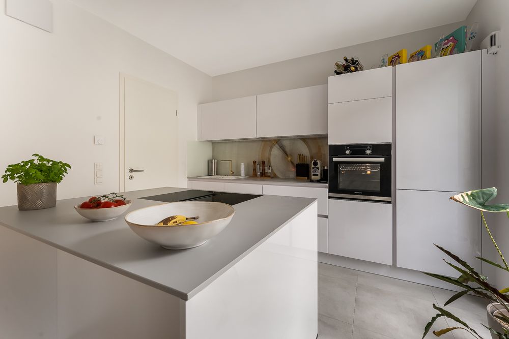 Moderne weiße Küche mit Kochinsel, Backofen und Schalen mit Obst und Kräutern auf der Arbeitsplatte.