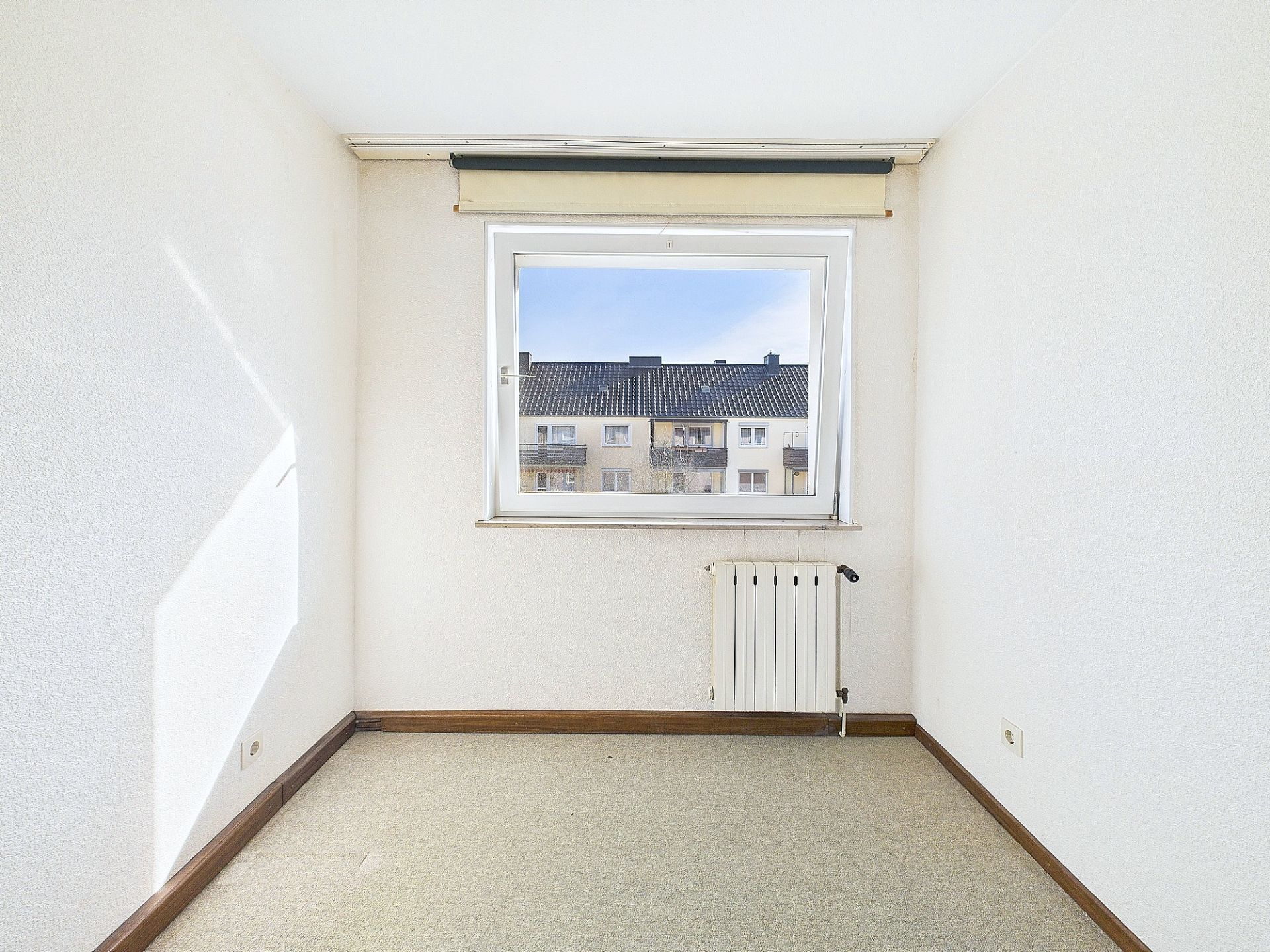 Kleiner, leerer Raum mit Fenster und Heizkörper, Blick auf benachbarte Wohnhäuser.