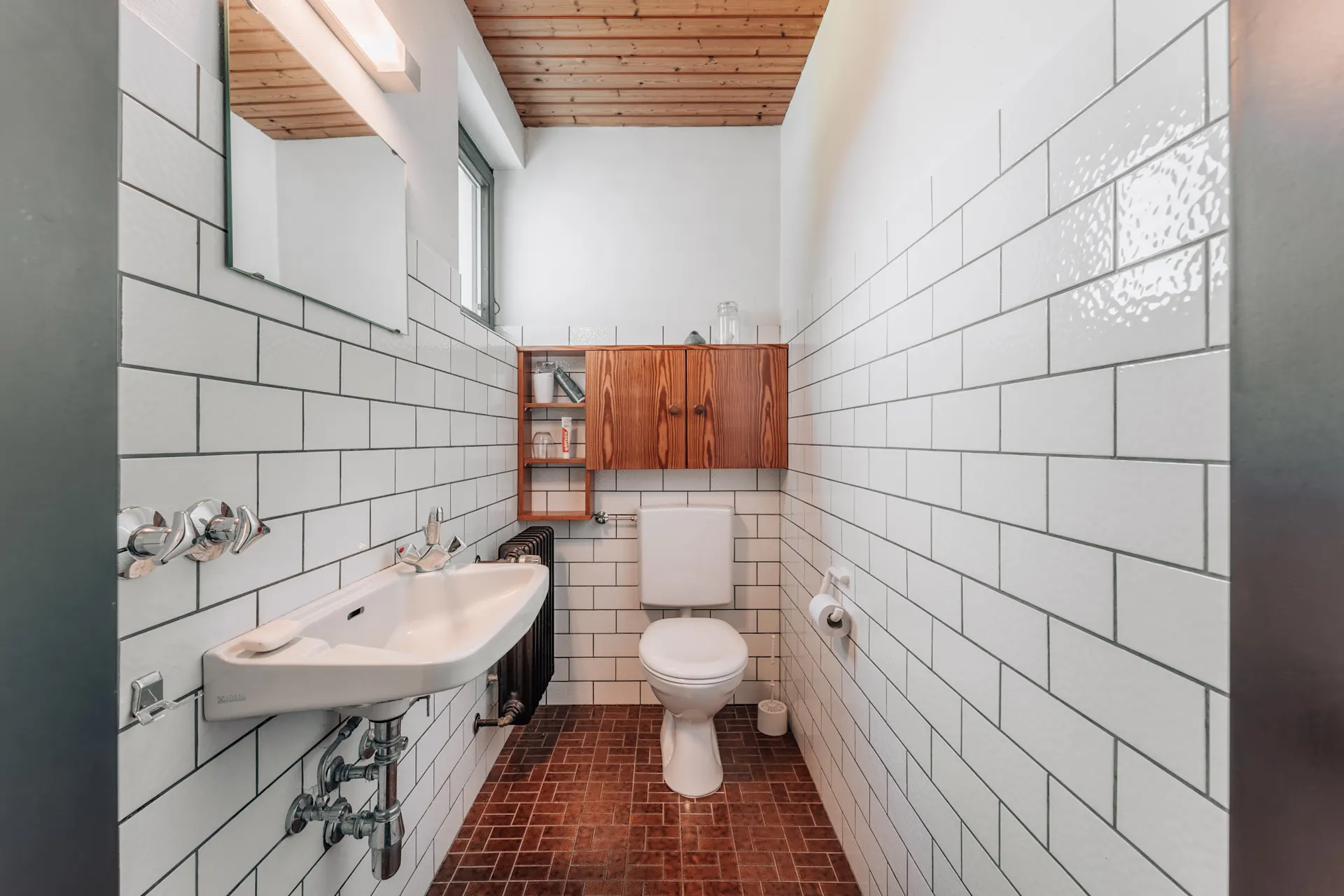 Helles Badezimmer mit weiß gefliesten Wänden, Waschbecken, Toilette und Holzschrank.