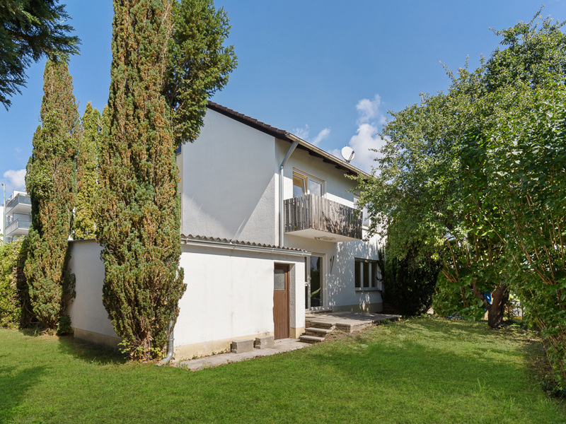 Zweistöckiges Wohnhaus mit Balkon und Garten, umgeben von Bäumen und Grünfläche.