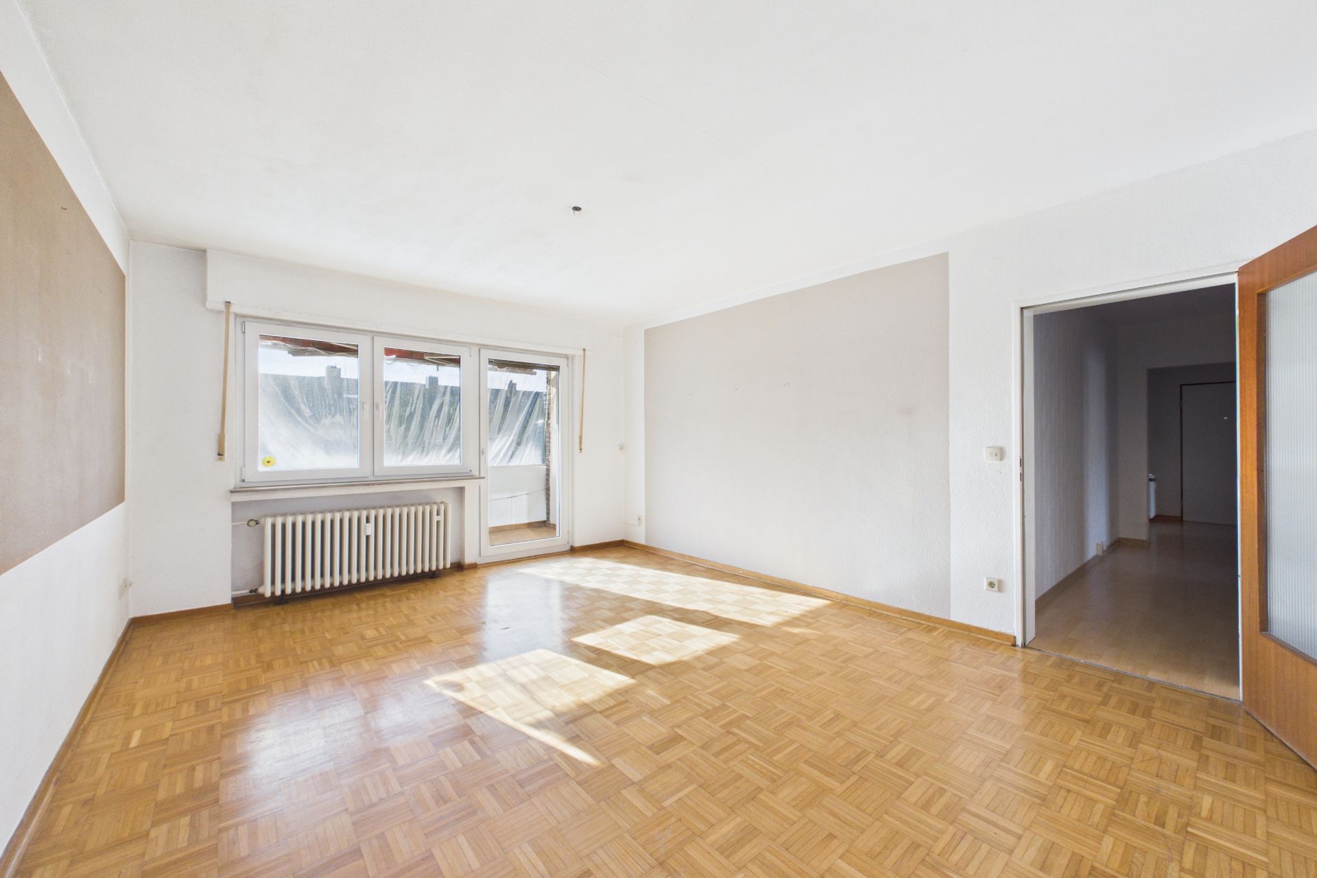 Helles, leeres Wohnzimmer mit Parkettboden, großen Fenstern und Zugang zu einem weiteren Raum.