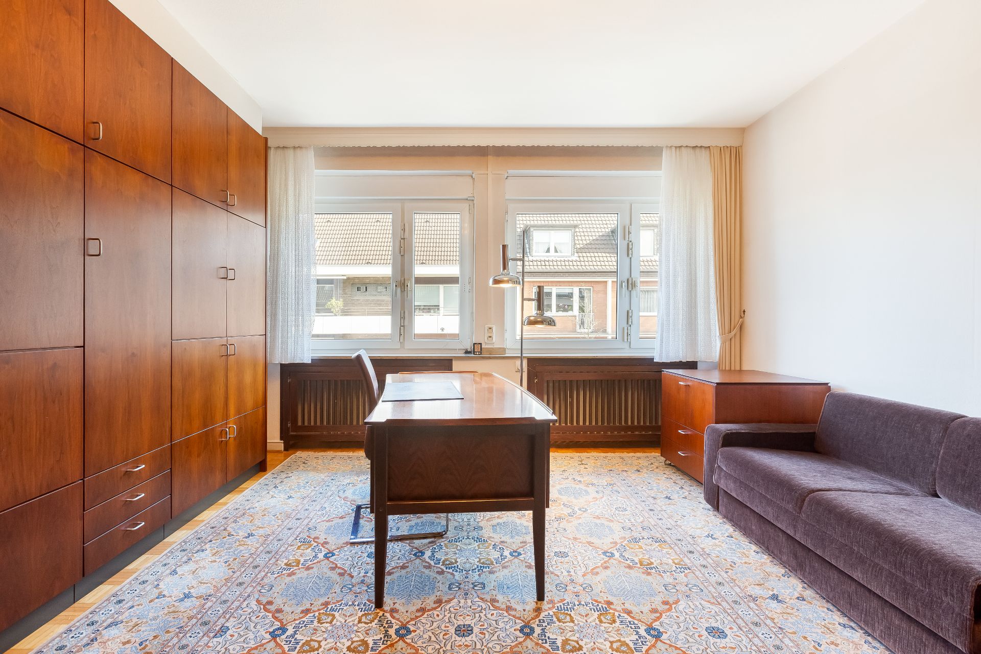 Helles Arbeitszimmer mit großem Schreibtisch, Teppich, Sofa und Holzschränken vor drei Fenstern.