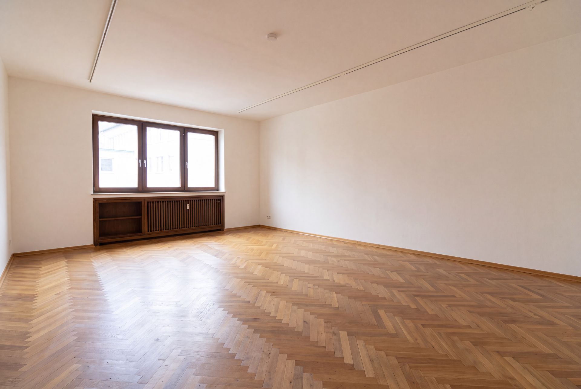 Leerer Raum mit Fischgrätparkett, großem Fenster und heller Wand.