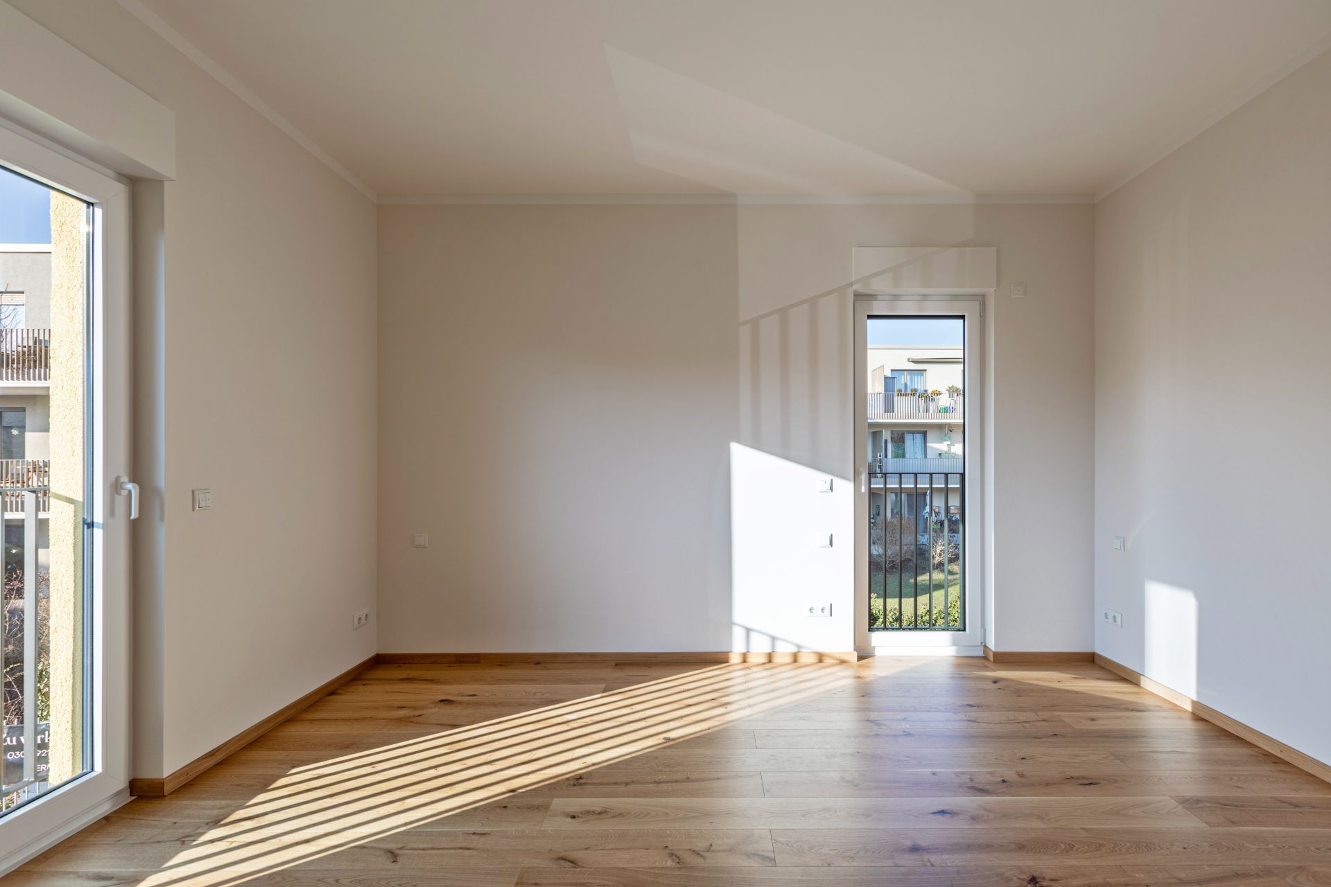 Helles, leeres Zimmer mit Holzfußboden und großen Fenstern, Sonnenlicht fällt herein.