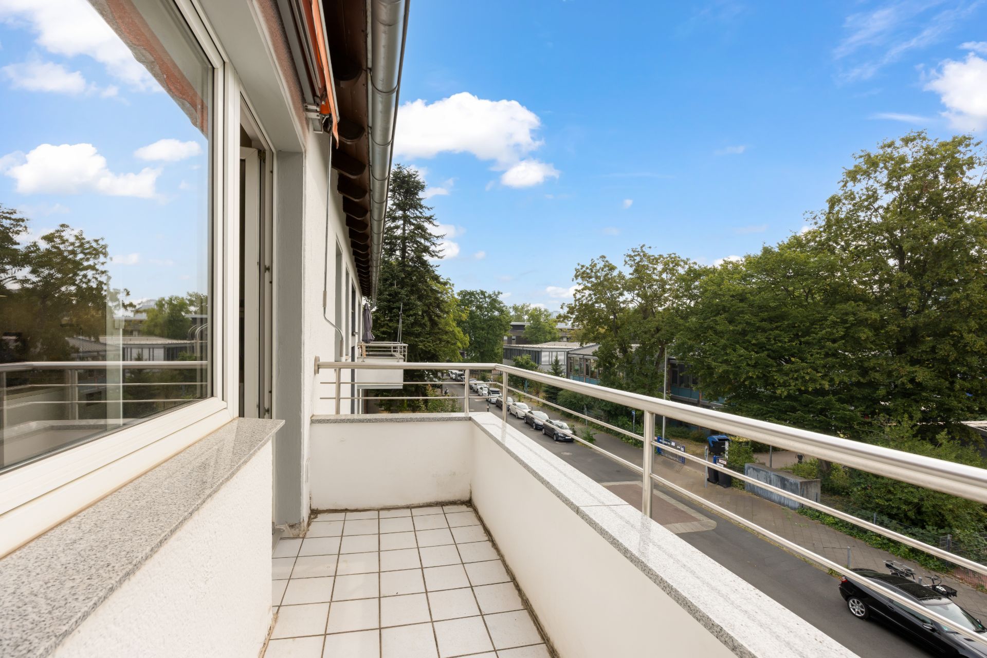 Balkon mit weißem Fliesenboden und Metallgeländer, Blick auf eine Straße mit Bäumen.