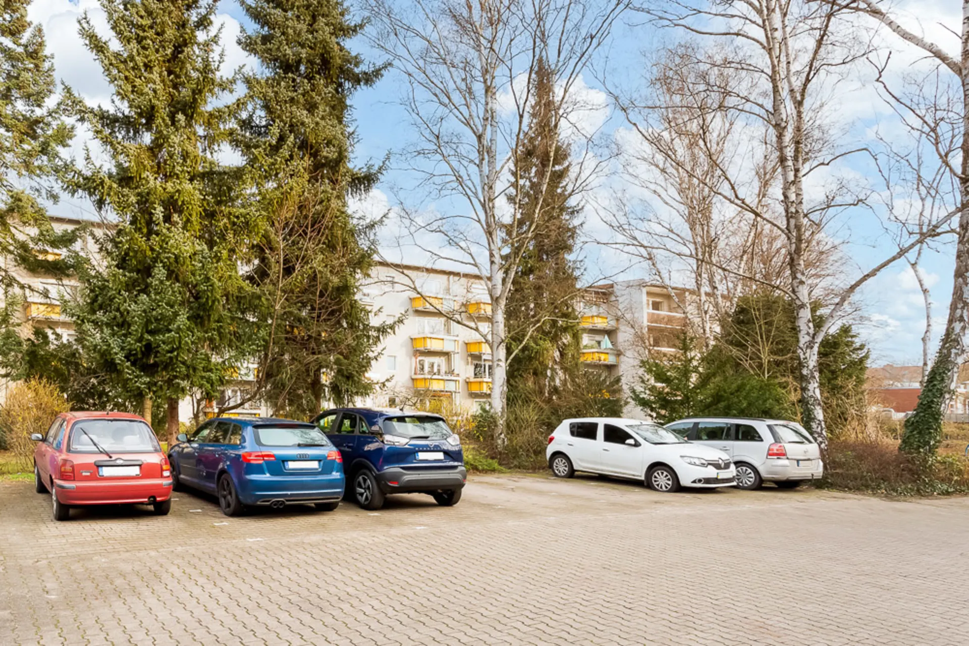 Mehrere Autos parken auf einem Parkplatz vor Wohnhäusern und Bäumen.