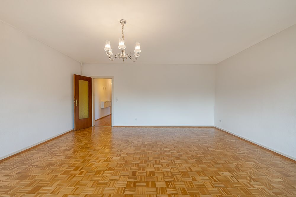 Leerer Raum mit Parkettboden, weiß gestrichenen Wänden und Kronleuchter, offene Tür im Hintergrund.