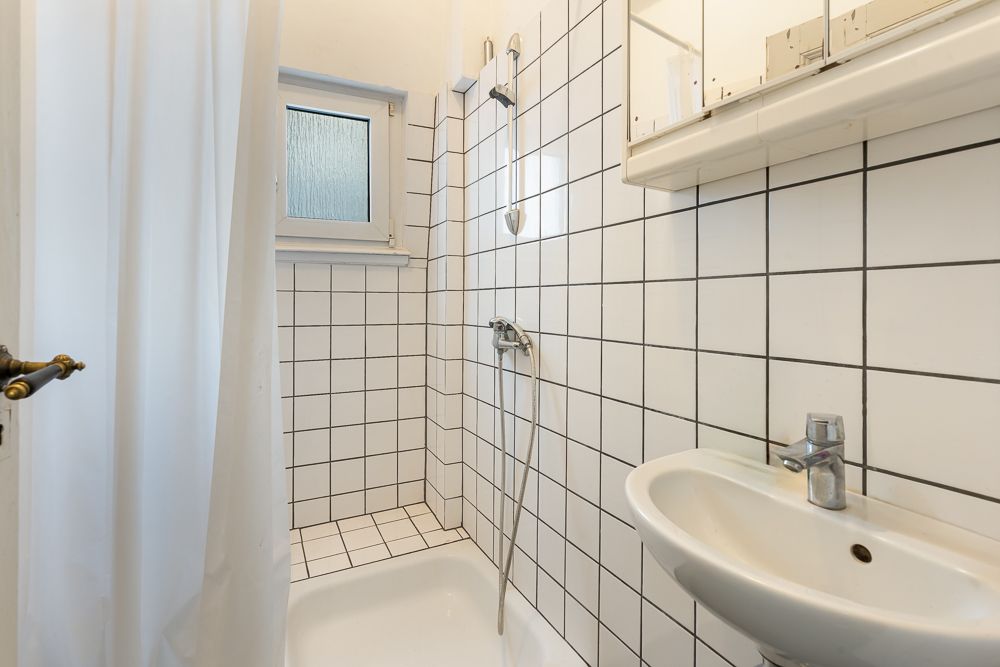 Kleines Badezimmer mit Dusche, Waschbecken, Fenster und weißen Fliesen an den Wänden.