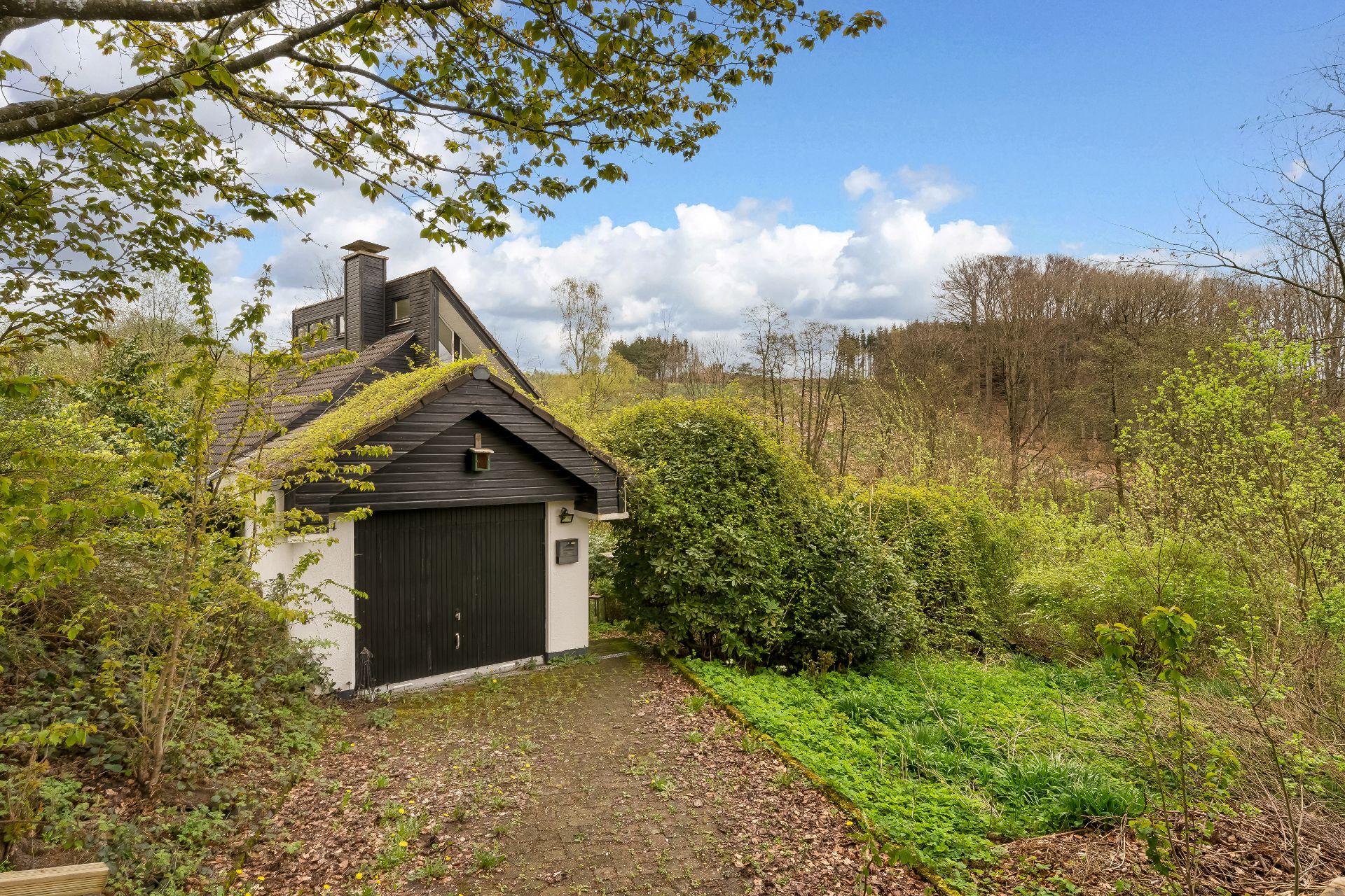 Haus mit schwarzem Garagentor und bewachsenem Garten, umgeben von Bäumen.